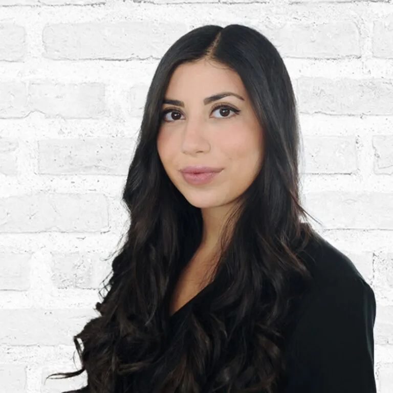 Portrait of a woman with long dark hair and a confident expression, standing against a white brick wall background at a plastic surgery center.