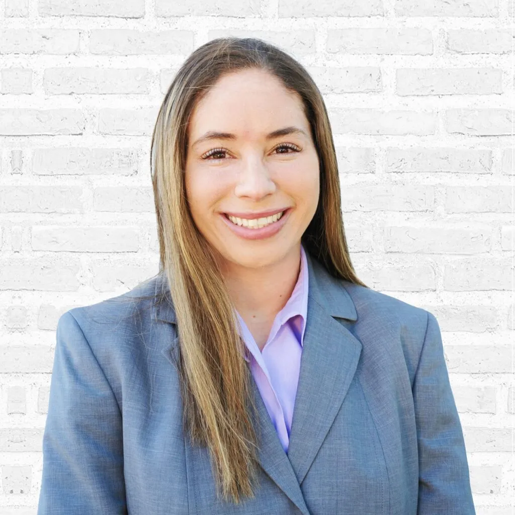 Smiling professional woman in a gray suit against a textured brick background.