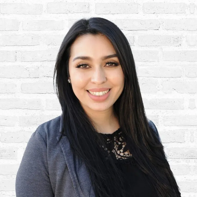 Smiling woman with dark hair in professional attire against a white brick background.