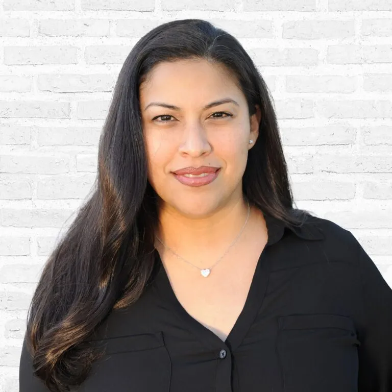 A person smiling in front of a white brick wall, wearing a black blouse and heart-shaped necklace.