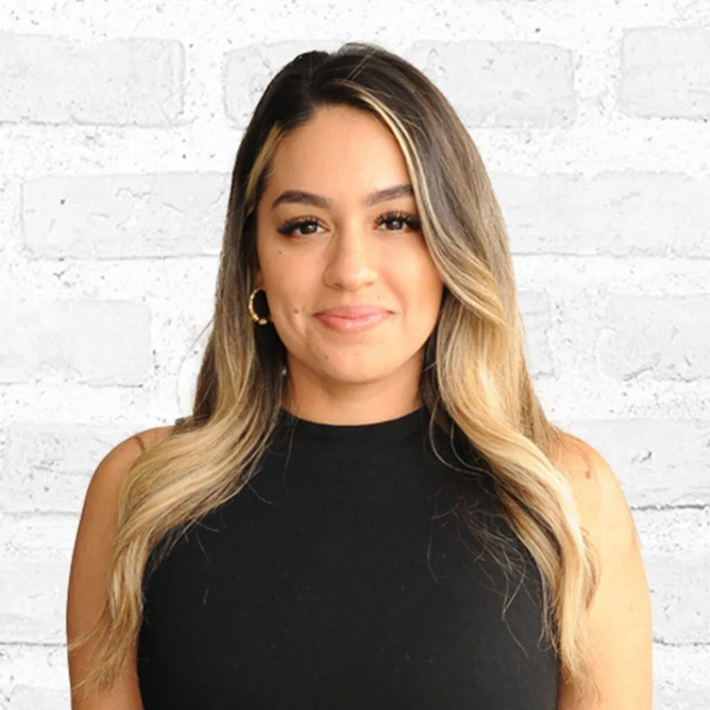 Woman with long, wavy hair smiling in front of a white brick wall.