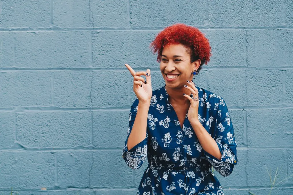 Smiling woman with curly red hair pointing, standing against a blue brick wall.