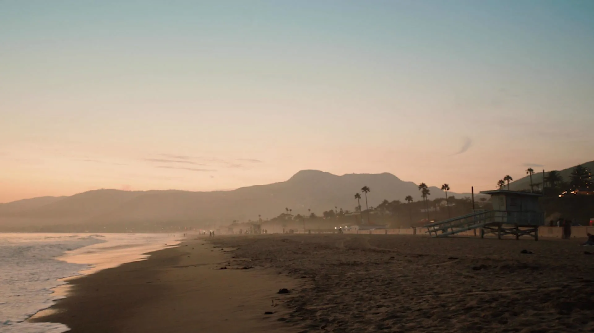 Scenic beachfront view at sunset with distant mountains and palm trees.