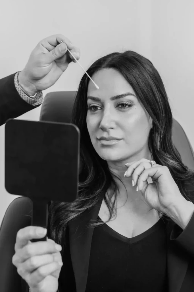 Woman contemplating cosmetic procedure while holding hand mirror, consultation at plastic surgery clinic.
