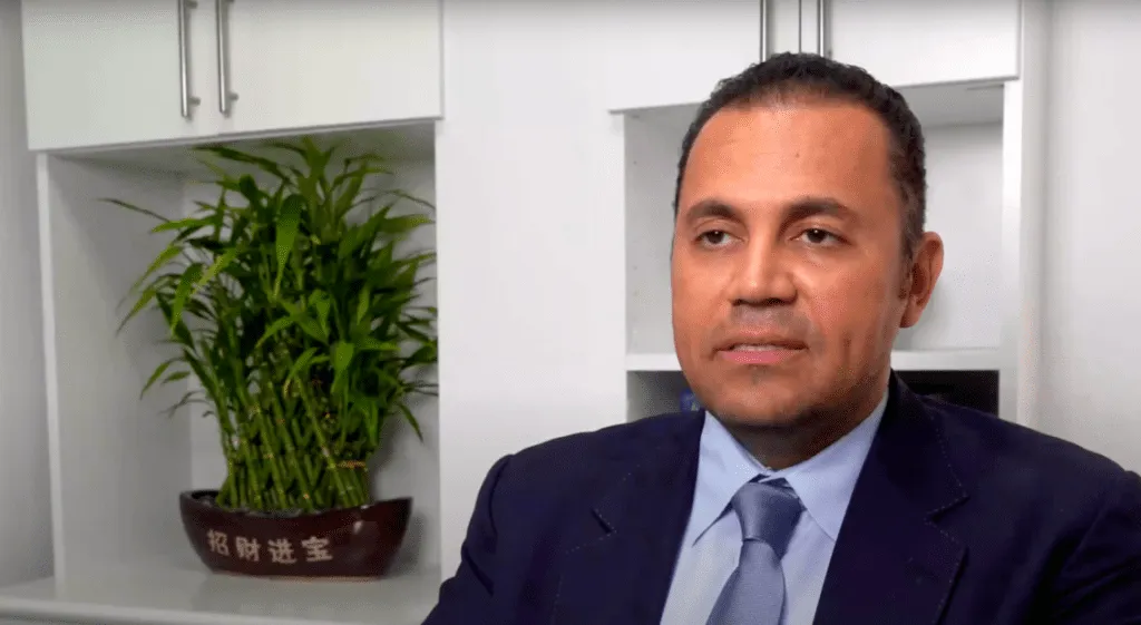 Doctor in a suit sitting in an office with a bamboo plant in the background.