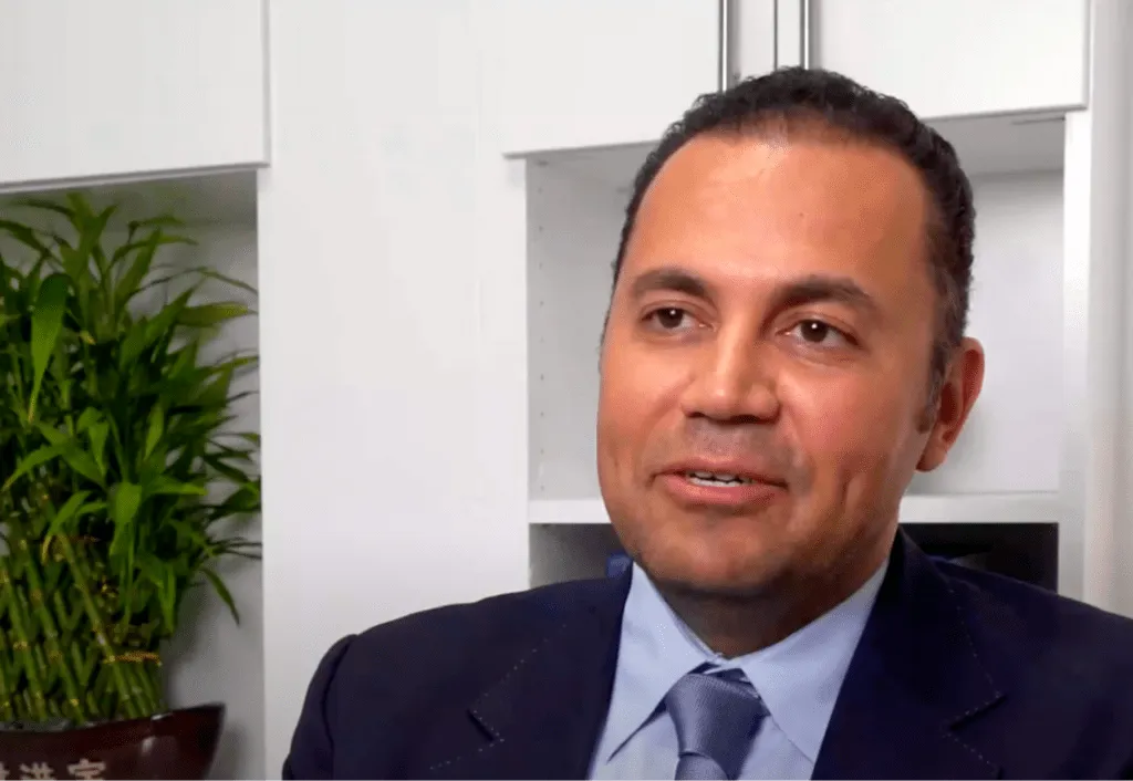 Smiling plastic surgeon in professional attire with plants in the background.