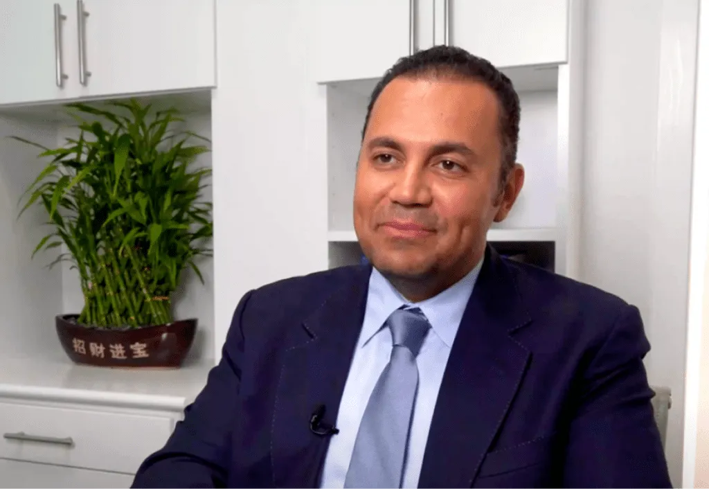 Man in a suit smiling with a bamboo plant in the background at a plastic surgery center office.