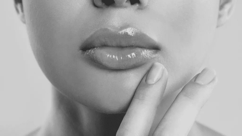 Black and white close-up of a person's lower face, focusing on full, glossy lips with manicured fingers resting lightly on the chin.