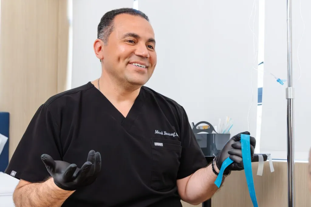 Smiling plastic surgeon in black scrubs holding medical tape in a clinic setting.