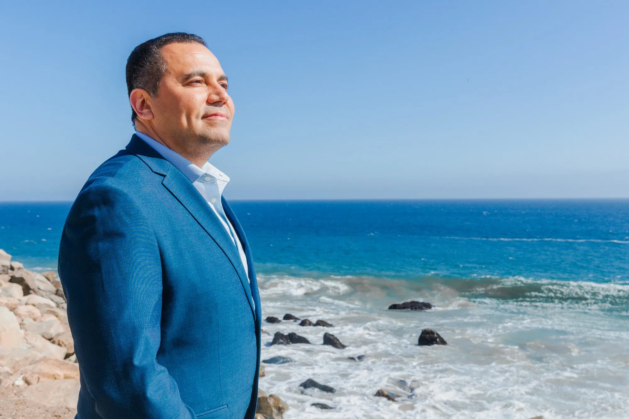 Man in a blue suit looking out over the ocean against a clear sky.