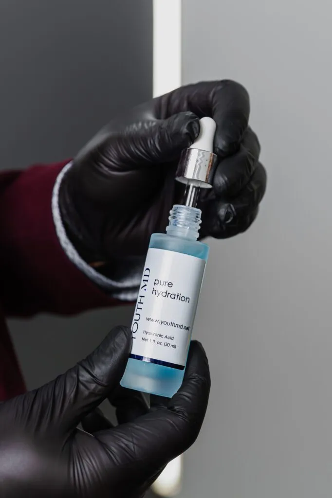 Gloved hands holding a bottle of hyaluronic acid serum with a dropper at a plastic surgery clinic.