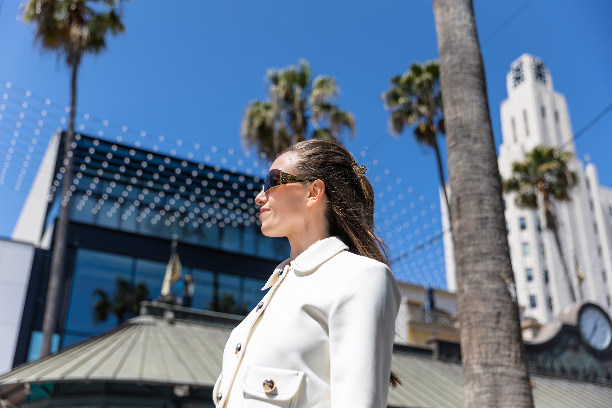 Woman in white blazer with sunglasses smiling outdoors against a blue sky.