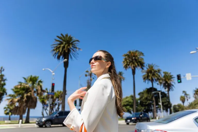 Woman in sunglasses and white blazer walking confidently in sunny outdoor setting with palm trees.