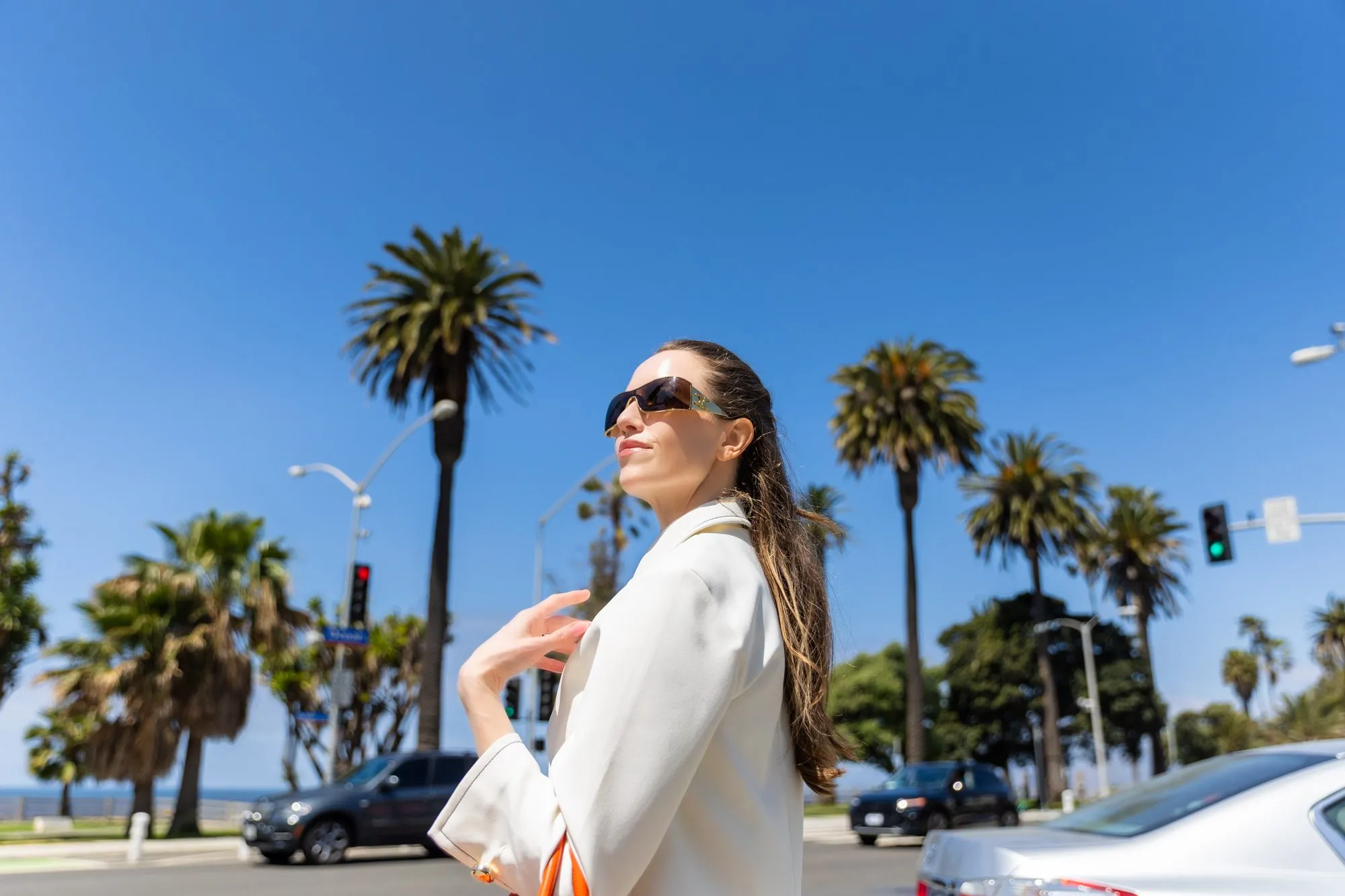 Woman in sunglasses and white blazer walking confidently in sunny outdoor setting with palm trees.